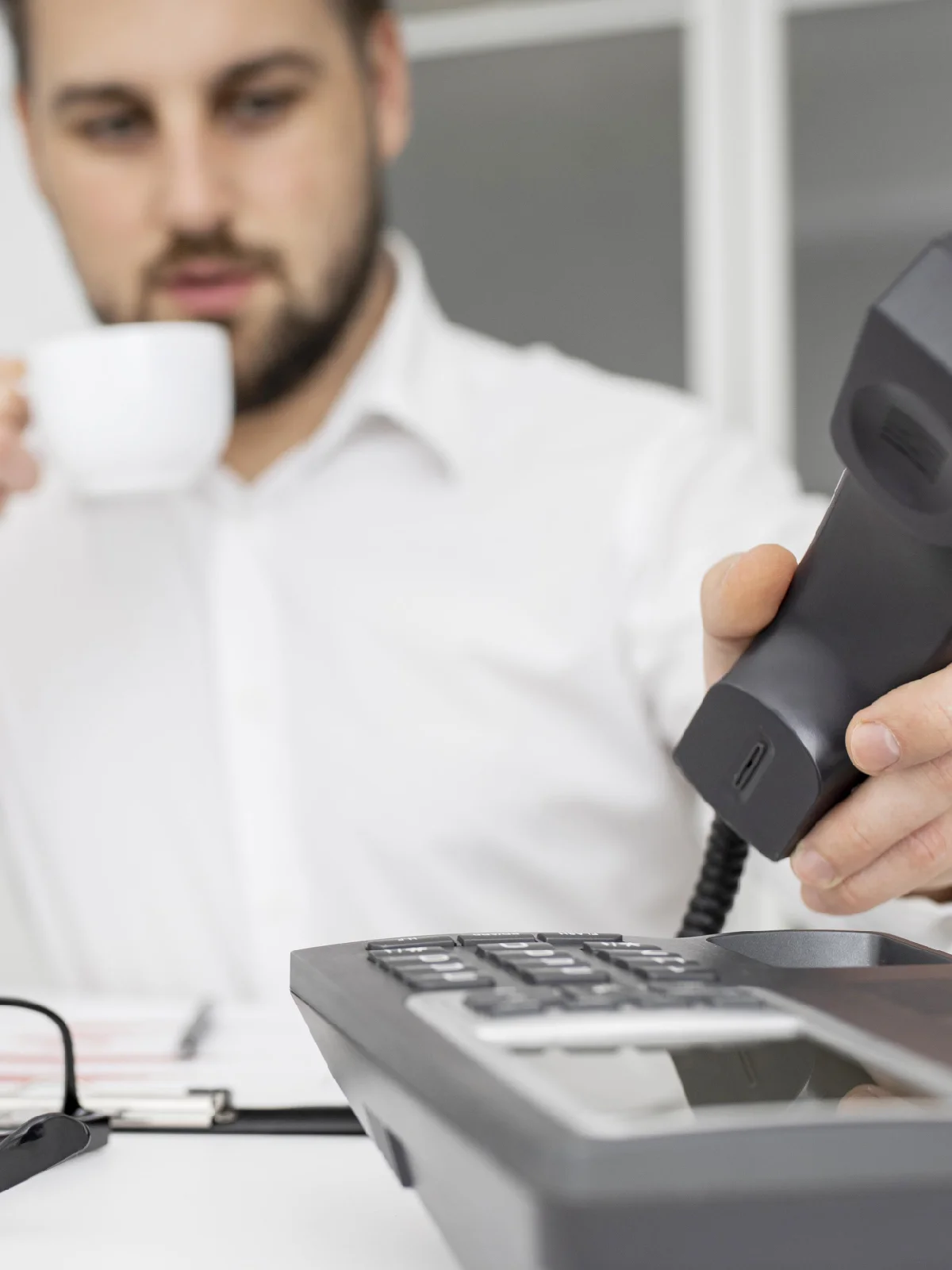 Office worker holding a coffee cup while reaching for a ringing desk phone